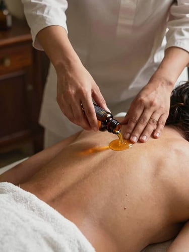 Close overhead shot of a Pakistani female therapist's hands pouring warm essential oil from a dark glass bottle over a client's upper back, amber oil catching warm studio light, white linen draped below, dark wood treatment table edge visible, intimate and precise