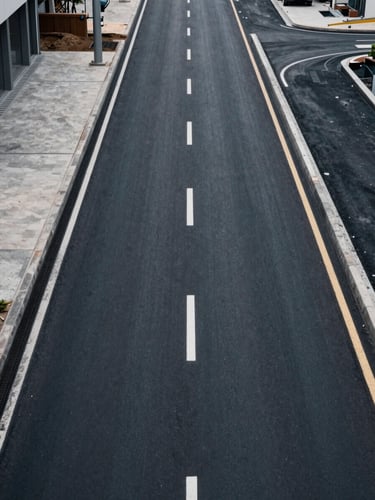 Overhead plan-view of a finished urban road corridor—freshly laid asphalt, clear lane markings, drainage infrastructure visible at margins, muted daylight, no vehicles or pedestrians, documenting completed deliverable scope from elevation