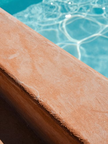 Extreme close-up of textured terracotta resin surface, sparkling pool water reflecting light in the background, bright direct sunlight, high-contrast shadows.