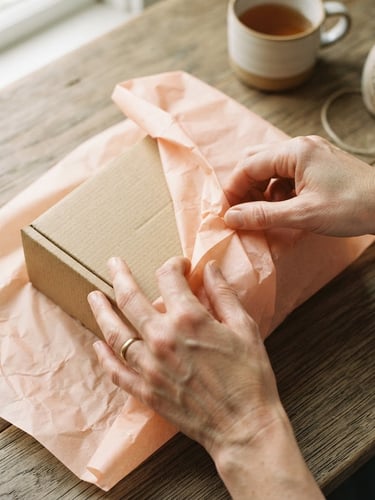 Overhead flat-lay of hands carefully wrapping a textured cardboard gift box in soft peach tissue paper, natural window light, muted organic tones, shallow depth of field