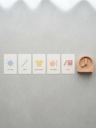 Overhead flat-lay of printed daily routine cards with simple icons, next to a clean wooden toy on a light gray surface, soft natural daylight, minimalist composition