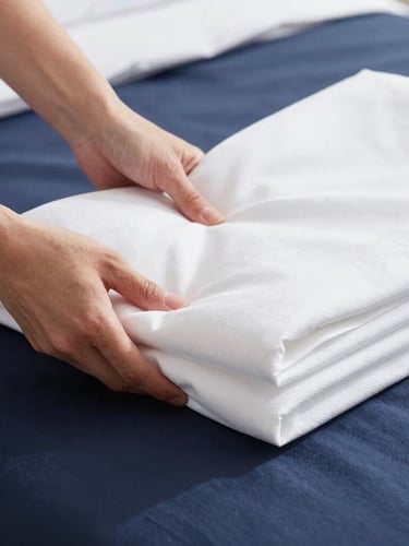 Bright commercial photography, crisp natural daylight, clean white and deep navy tones, sharp focus, 35mm lens, close-up of precise hands folding crisp white hotel linens on a bed