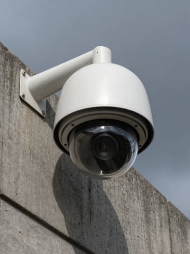 Low-angle sharp shot of a permanent commercial security camera dome mounted on a concrete industrial building corner, dark overcast sky, sharp steel and concrete textures, gritty realistic lighting.