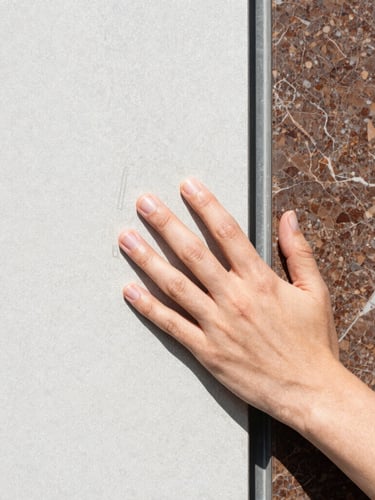 Close-up detail of hands touching a textured sheet of Tyvek next to a piece of polished brown Emperador marble, bright natural daylight, high-contrast editorial