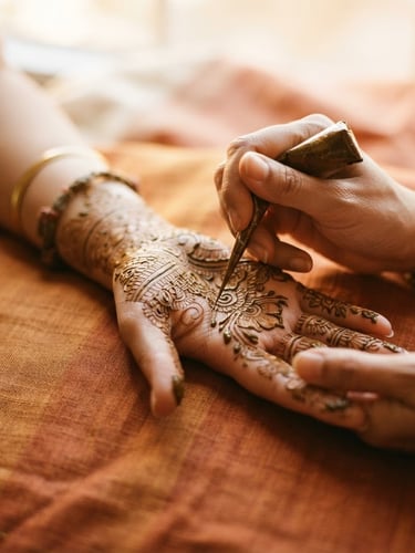 Close-up of hands resting on raw silk, artist applying intricate mehendi lines with a natural henna cone, warm diffused window light, shallow depth of field, 35mm