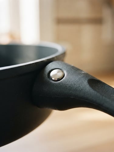 Close-up detail of a matte non-stick frying pan handle, stainless steel rivet, soft morning window light, warm neutral background.