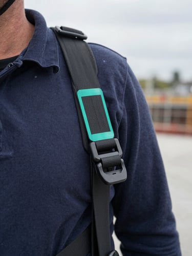 Close-up side angle of a construction worker's torso and shoulder, a safety harness strap partially unclipped, a faint teal bounding-box overlay highlighting the unclipped buckle, overcast natural light on a concrete site, no faces visible