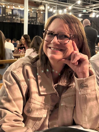 Smiling woman with glasses wearing a tan corduroy jacket at a dinner event with string lights.
