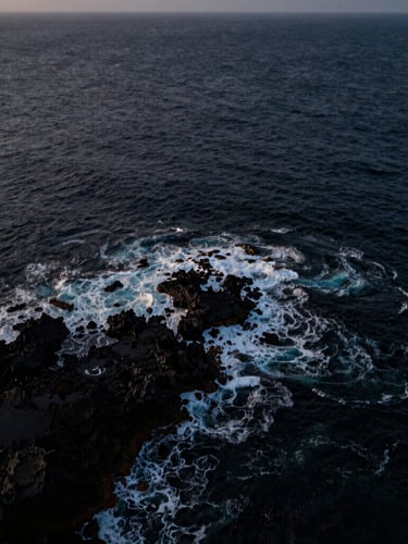 Wide panoramic aerial shot of a rugged coastline at blue hour, deep dark ocean waves crashing against black volcanic rocks, captured from a high-altitude drone pass, moody cinematic lighting.
