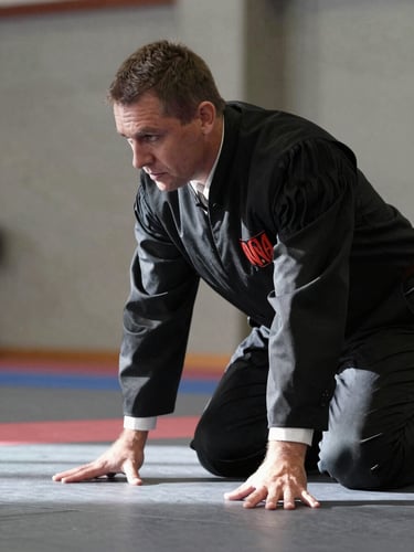 Torso-level framing inside a battle venue, a judge leaning forward over the floor with arms resting on knees, face in sharp concentration, side-angle strobe light casting hard shadows across their jacket and hands, muted concrete walls in background, no crowd visible