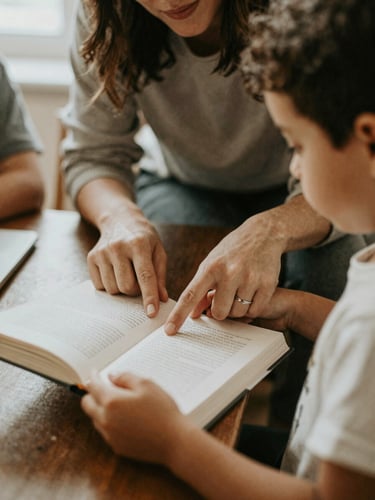 Close-up of hands-on collaborative work, a teacher guiding a child pointing to a book, soft morning light, warm editorial style, muted earth tones.