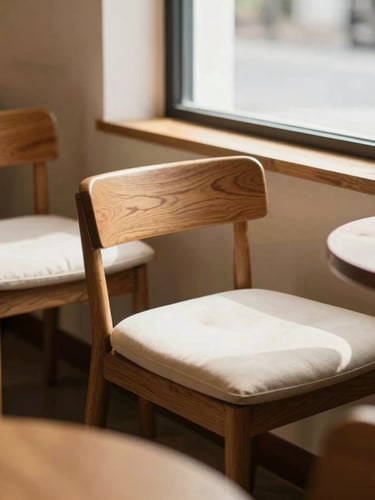 A warm editorial photograph of a cozy café corner, soft natural window light streaming across comfortable wooden chairs and cream cushions, shallow depth of field