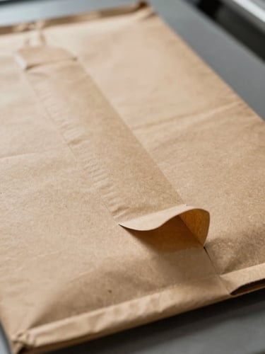 Extreme close-up of a kraft paper mailer laid flat on a warehouse sorting surface, both adhesive strips visible — the first strip sealed and the second strip tab folded open, sharp studio lighting from above, mailer texture crisp and detailed