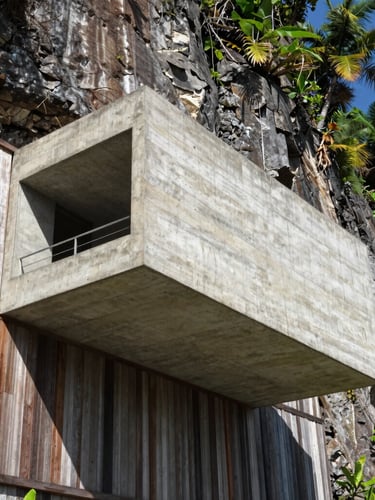 Low-angle architectural photograph of a raw concrete cantilevered volume over a steep tropical cliff, harsh midday light casting sharp diagonal shadows, weathered teak wood screens, 35mm lens.