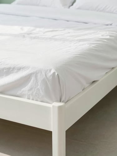 Straight-on architectural photo of a perfectly made bed with crisp white hospital corners, bright high-key natural daylight, clean white and soft green tones, sharp focus, 35mm