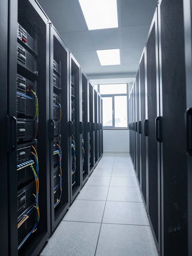 Wide environmental shot of a server room corridor under overhead natural light, rows of rack-mounted equipment receding into depth, no people, cables neatly dressed along the rails, cool daylight spilling through a high window on the right side