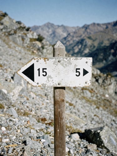 Close-up of a weathered trail marker on a rugged mountain pass, harsh high-altitude daylight, cold shadows, desaturated alpine tones