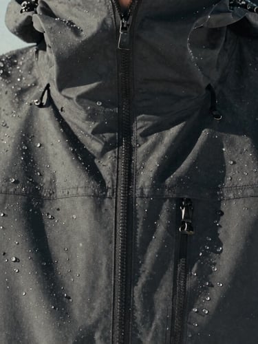 Close-up of a technical hardshell jacket covered in ice droplets, alpine environment, harsh high-contrast natural light, desaturated tones