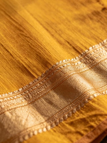 Macro detail shot of a warm turmeric hand-spun cotton saree border, soft golden-hour sunlight highlighting the raw textile texture, shallow depth of field.
