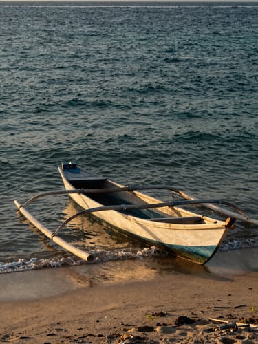 A traditional wooden fishing boat (perahu nelayan) resting on the sandy shores of Kuala Batee, Southwest Aceh, captured during golden hour with warm sunlight reflecting on the gentle waves.