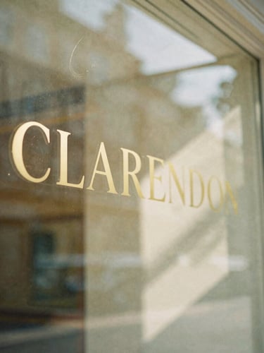 Intimate close-up of a sunlit Clarendon storefront window with gold lettering, soft morning light reflecting on glass, warm shadows, 35mm film look