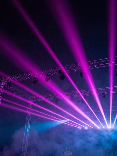Low-angle shot of massive aluminum stage truss rigging silhouetted against intense magenta laser beams, smoke machine haze, 35mm dramatic photography
