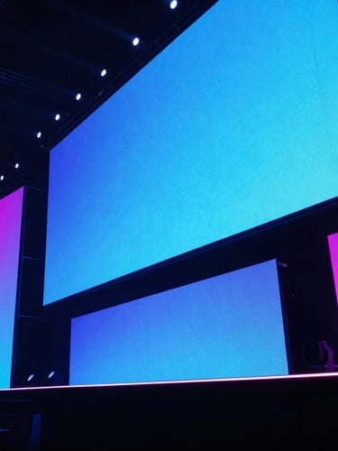 Low-angle dramatic shot of a massive corporate stage, sharp neon blue and magenta backlighting, crisp LED screens, ambient venue glow, 35mm