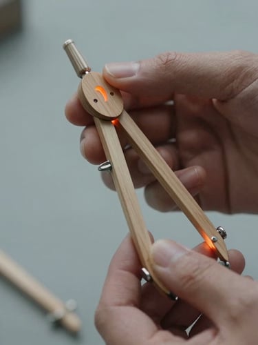 Tactile close-up of hands holding a wooden compass prototype with soft orange glowing light, shallow depth of field, cool grey background