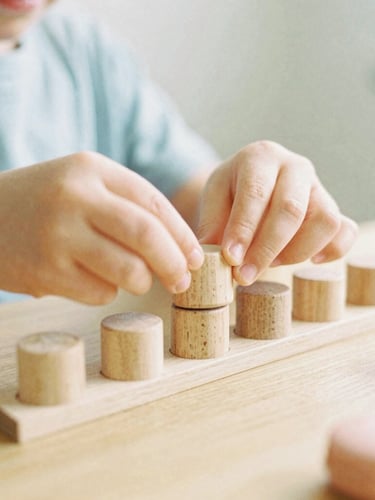 Candid eye-level shot of a child's hands carefully placing natural wooden cylinders into a matching block, soft morning daylight, shallow depth of field, 35mm film style.