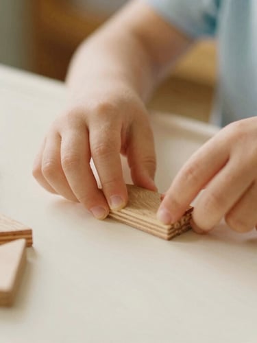 Close-up of a child's hands carefully arranging wooden geometric shapes on a low cream-colored table, warm morning daylight streaming in, shallow depth of field, soft pastel tones, 35mm film style.