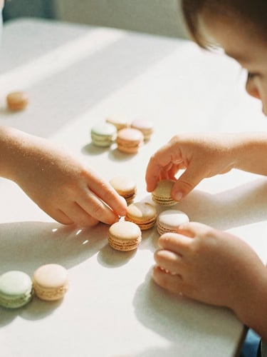 Warm editorial photography of a young child's hands sorting smooth wooden Montessori beads on a sunlit table, soft morning daylight, gentle macaron pastel tones, shallow depth of field, candid, 35mm film style