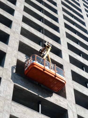 Low-angle architectural shot of a safety-orange passenger hoist climbing a modern concrete skyscraper under bright daylight, 35mm