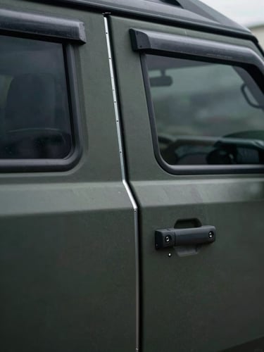 Close-up exterior detail of an armored SUV door panel, ballistic-rated glass edge visible, steel reinforcement seam sharp under cool directional daylight, dark matte finish, no people, industrial precision framing