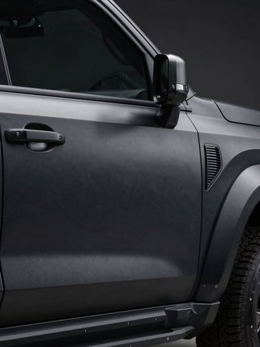 Close-up exterior panel of a matte black armored SUV, right-side framing, studio strobe lighting raking across the steel bodywork to reveal paint texture and door seam precision, extreme detail on the B-pillar and ballistic-rated window edge, dark background, no people