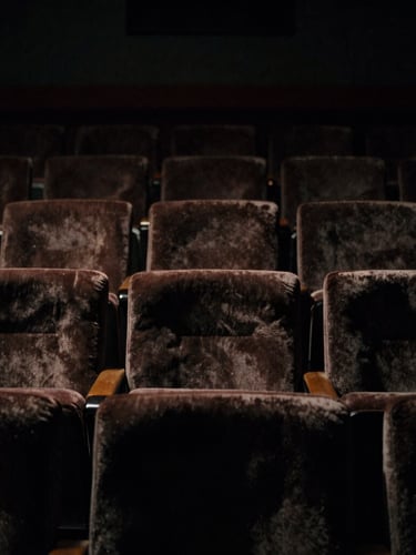 Dramatic low-key cinematic shot of a dark theater interior, faint light catching the edges of empty velvet seats, 35mm anamorphic lens style.
