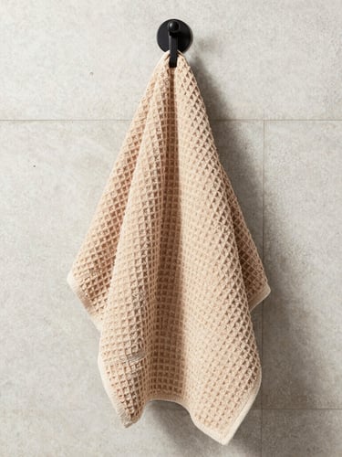 Straight-on architectural crop of a warm oatmeal waffle-weave towel hanging from a matte black hook on a light concrete tiled wall, bright indirect natural daylight, sharp macro detail
