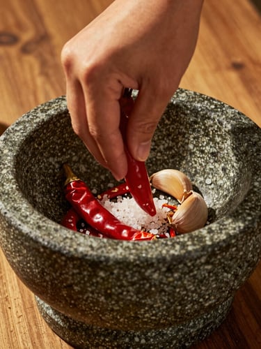 Close-up of a stone mortar with fresh red chilies, garlic, and sea salt being ground by hand, warm golden hour lighting, rustic wooden table