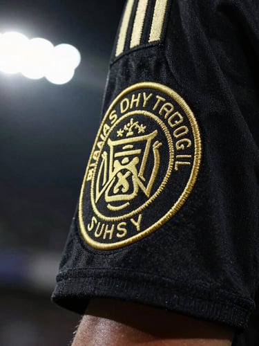 Dramatic close-up of a premium football jersey sleeve, showcasing high-contrast gold embroidered crest stitching, deep black fabric texture, stadium floodlight illumination, 35mm