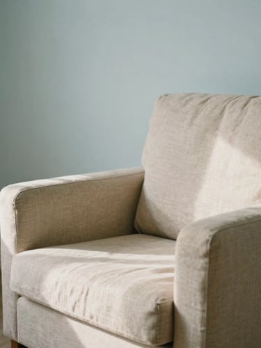 Soft environmental shot of an empty therapy room with a comfortable armchair, linen textures, gentle morning shadows, muted sand and light blue tones, 35mm