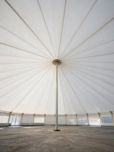 Wide environmental shot looking up at the interior of a large white PVC big-top canopy, radial tension lines converging at the crown, daylight filtering through the fabric, empty interior, no people