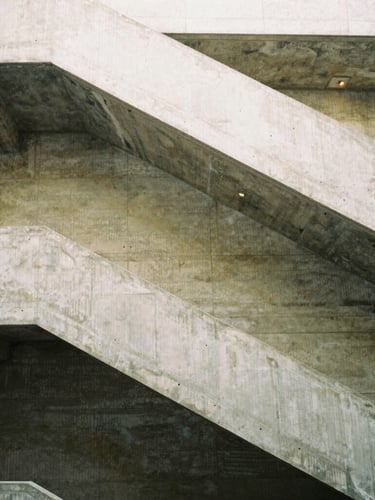 A tall vertical portrait of a concrete staircase winding upward, sharp architectural lines, soft diffused daylight falling from above, medium-format film style.