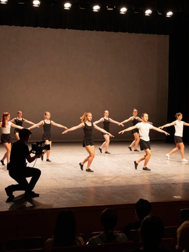 Wide environmental shot of a dance recital in progress, camera operator crouching at stage-left with a handheld rig, warm stage lighting on young dancers mid-performance, audience silhouettes visible in lower frame, practical tungsten from overhead
