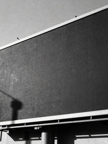 High-contrast black and white photography of a massive physical billboard in a stark urban landscape, harsh direct sunlight, deep dramatic shadows, 35mm film grain