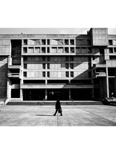 Wide panoramic shot of a brutalist concrete building facade in harsh direct sunlight, casting dramatic, sharp shadows. A single figure in silhouette walks across the foreground, creating a sense of scale and purpose. High-contrast black and white photography, sharp focus, 35mm film grain.