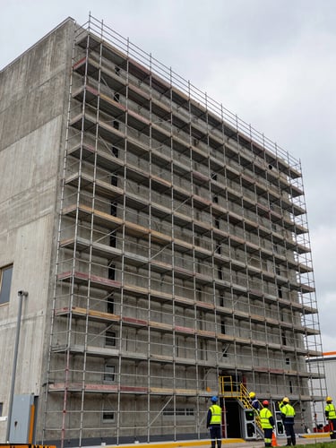 Wide environmental shot of a large industrial facility facade, scaffolding system installed against the building exterior in natural overcast daylight, high-visibility safety equipment visible, concrete surface texture and repair work in progress, building scale emphasized