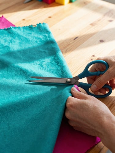 A woman hand-cutting colorful teal and magenta fabrics with scissors on a large wooden workshop table, warm natural sunlight filtering in, close-up details.
