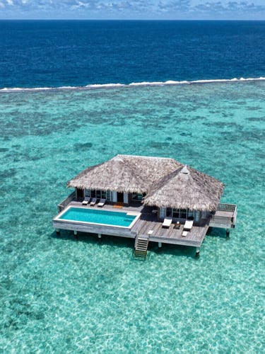 Wide aerial view of a single luxury villa on a private wooden deck over turquoise water in Bora Bora, sharp midday sun, endless deep blue ocean in the background, no people.