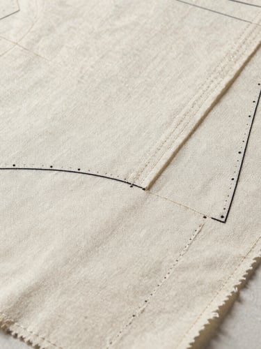 Diffused studio light on a technical garment pattern overlaying a heavy canvas fabric swatch, showing precise stitching details.