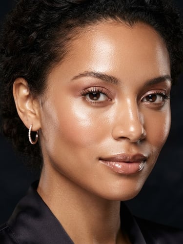 Editorial portrait of a diverse model with luminous, dewy skin, soft rose gold makeup tones, warm studio strobe lighting, high-contrast close-up crop, dark obsidian background.
