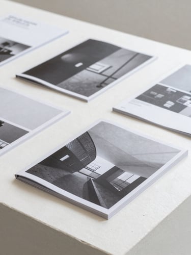 A desaturated flat-lay of printed contemporary exhibition catalogues and research journals on a white concrete table, soft natural overhead light, minimal composition